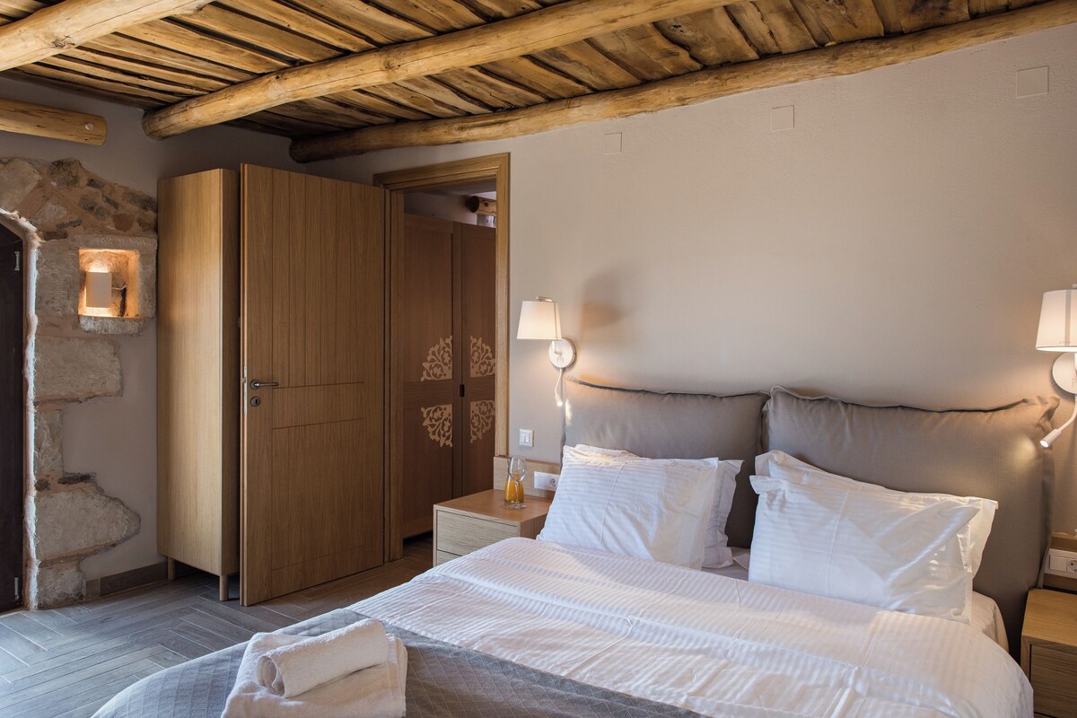 Une chambre avec un plafond en bois, un mur en pierre, une armoire en bois, une table de chevet avec une lampe et un lit avec du linge de lit blanc.