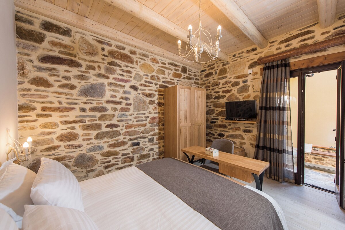 Une chambre avec un mur en pierre, des poutres apparentes en bois au plafond, un lustre, un lit, un bureau et un téléviseur.