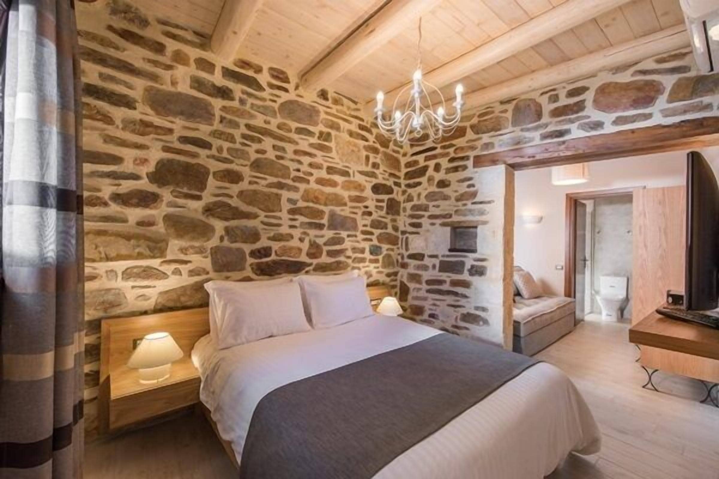 Un dormitorio con pared de piedra, una cama con ropa de cama blanca, una mesita de noche de madera con una lámpara y una araña.