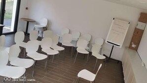 Ein Konferenzraum mit weißen Stühlen, die in Reihen angeordnet sind, einer Whiteboard-Tafel und einem kleinen Tisch mit Sitzgelegenheiten.