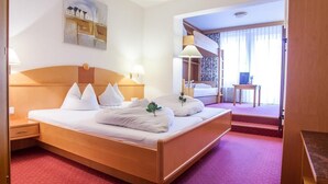A hotel room with a large bed, a bedside table, a lamp, a painting on the wall, and a view of another room through a door. - Auderer Hotel (Karroesten)