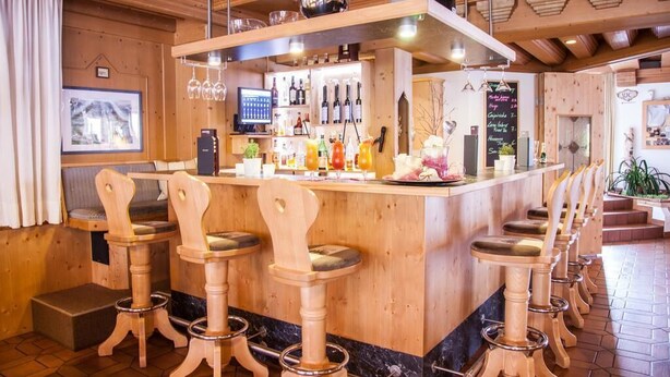 A bar area with wooden stools, a bar counter, and a variety of bottles and glasses. - Auderer Hotel (Karroesten)