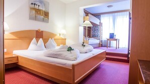 A hotel room with a large bed, a bedside table, a lamp, a painting on the wall, and a view of another room through a door. - Auderer Hotel (Karroesten)