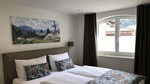 A hotel room with a large bed, a painting of a mountainous landscape, a window with curtains, and a view of a building outside. - Alpenschlössl (Mayrhofen)