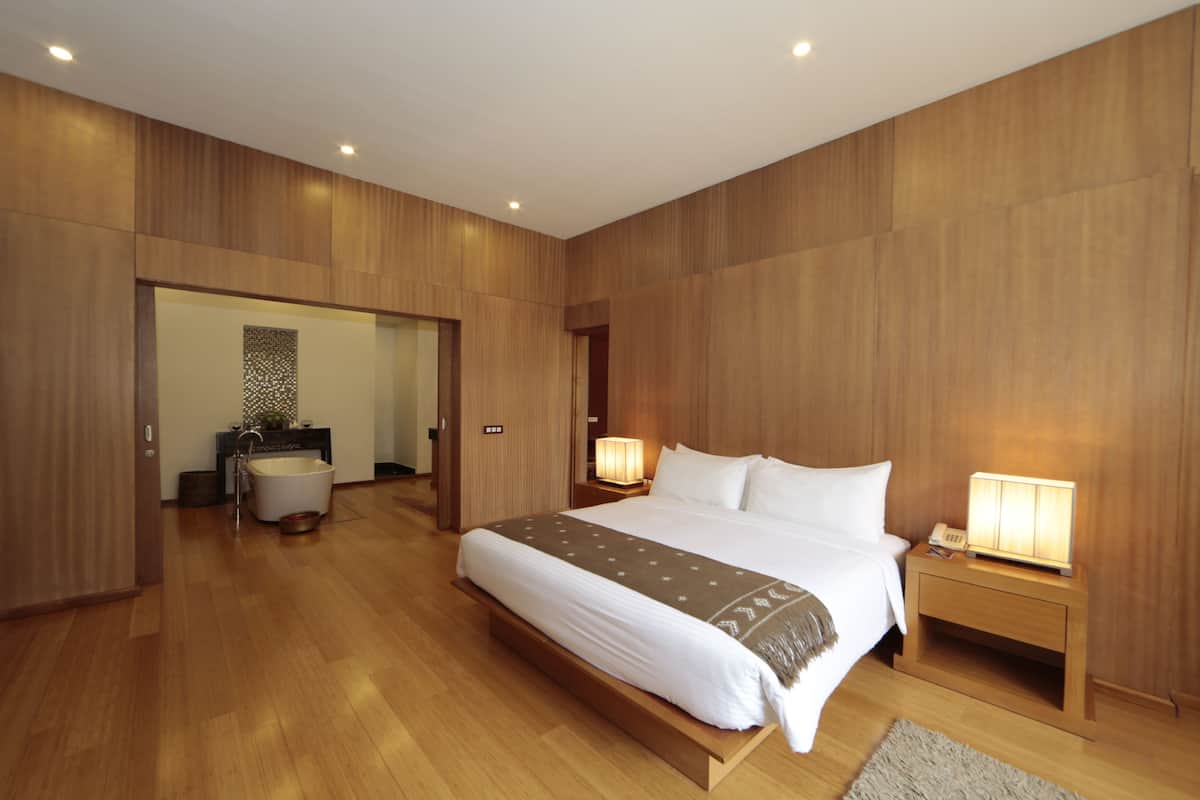 A modern hotel room with a large bed, wooden paneling, two bedside lamps, a nightstand, and a bathroom visible through an open door.