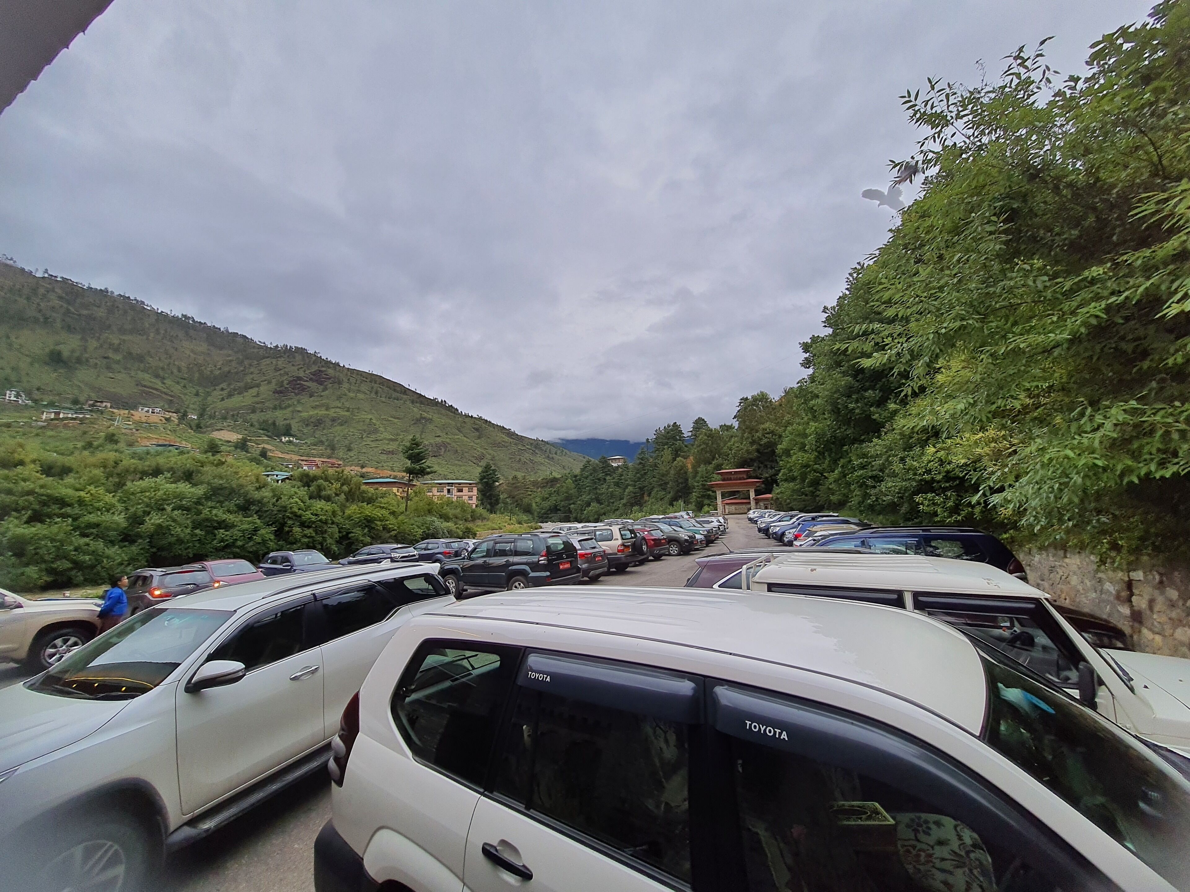 A parking lot with multiple cars, surrounded by lush greenery and hills in the background.