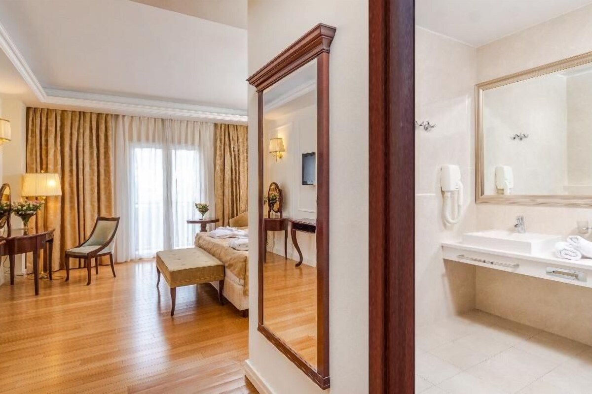 Habitación de hotel con piso de madera, cama, zona de estar con una silla y una mesa, un espejo grande y un baño con bañera y lavamanos.