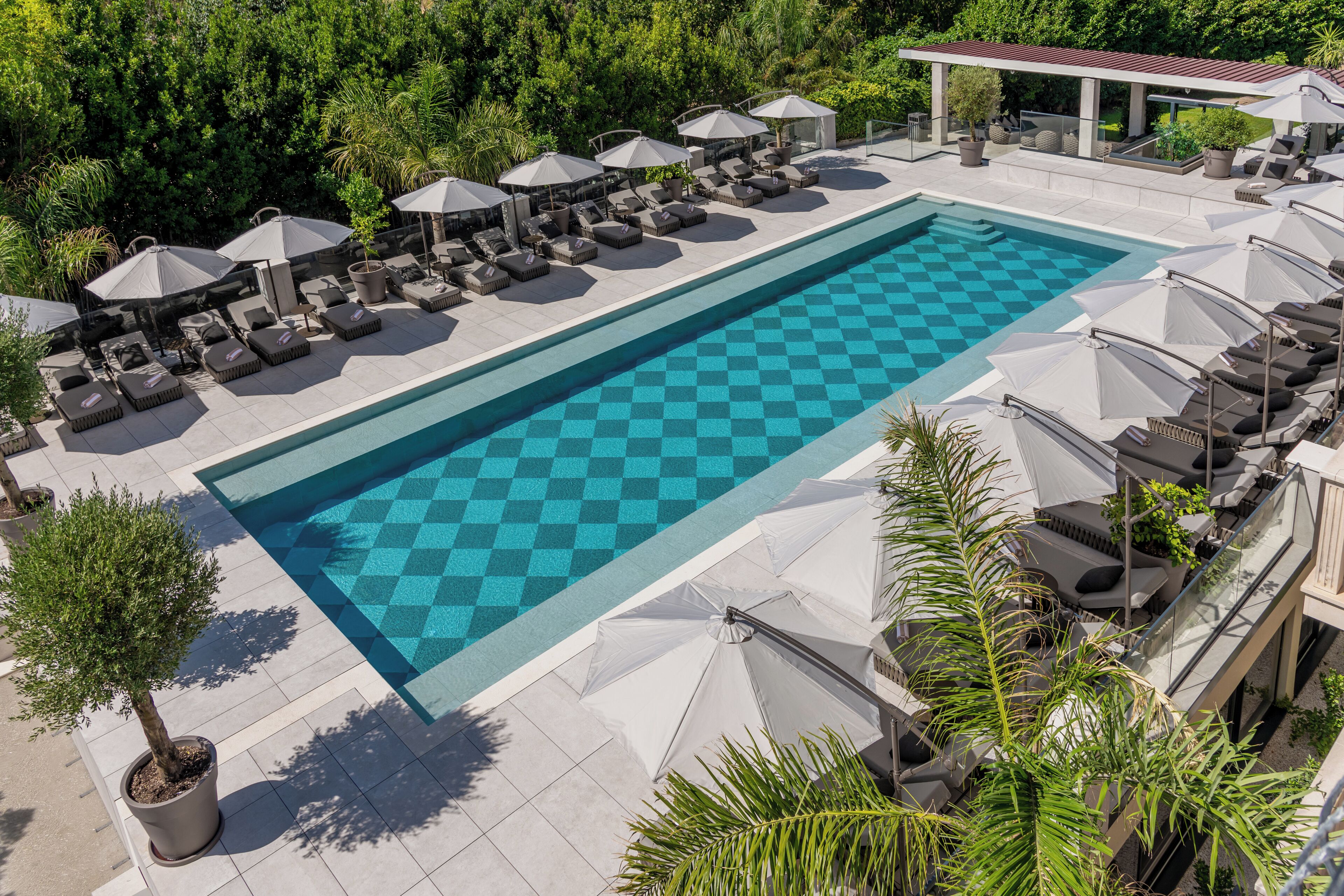A pool with checkered tiles, surrounded by lounge chairs and umbrellas, with a view of greenery and a covered area in the background.