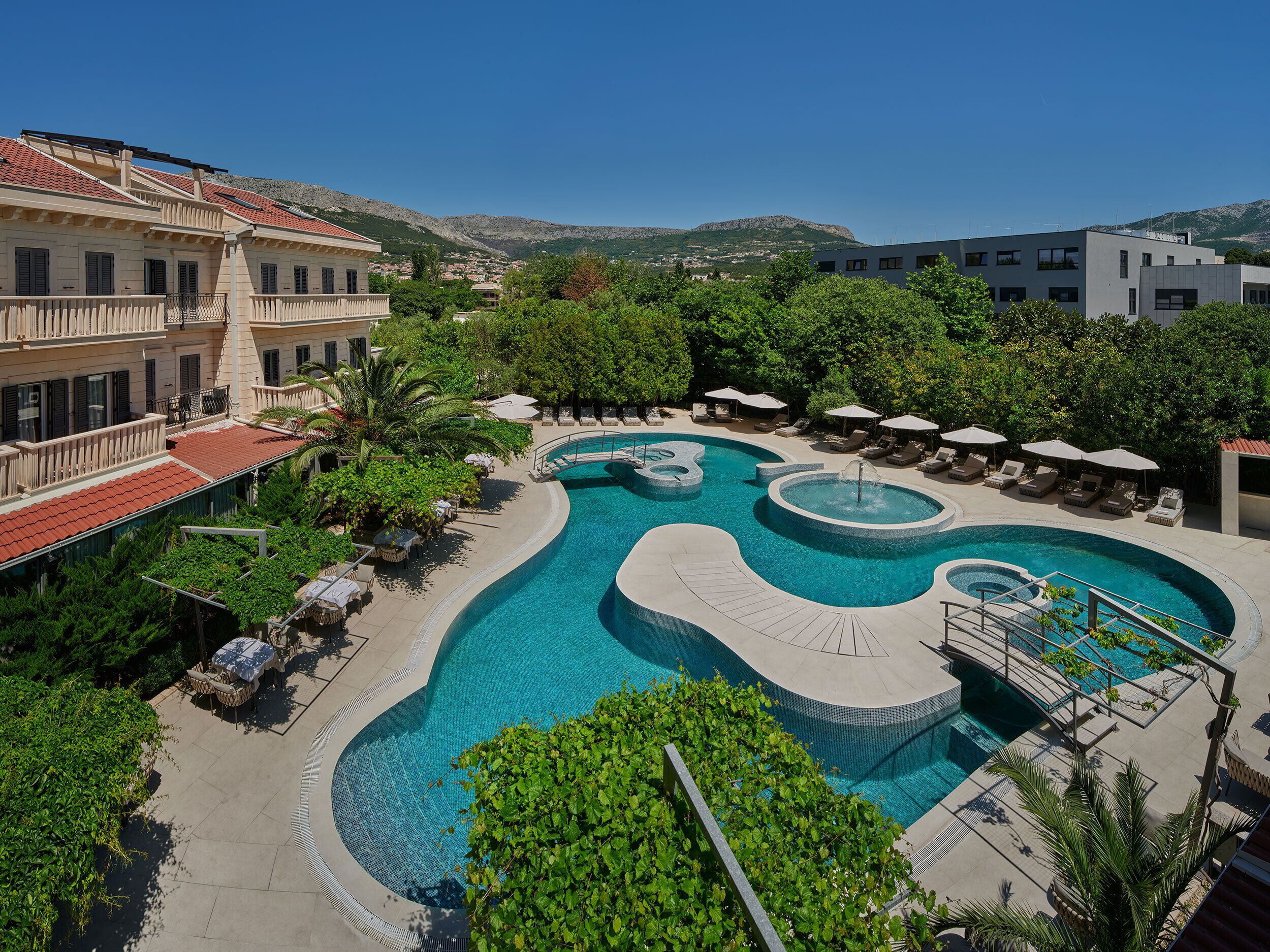 A swimming pool with multiple sections, surrounded by lounge chairs and umbrellas, with buildings and mountains in the background.