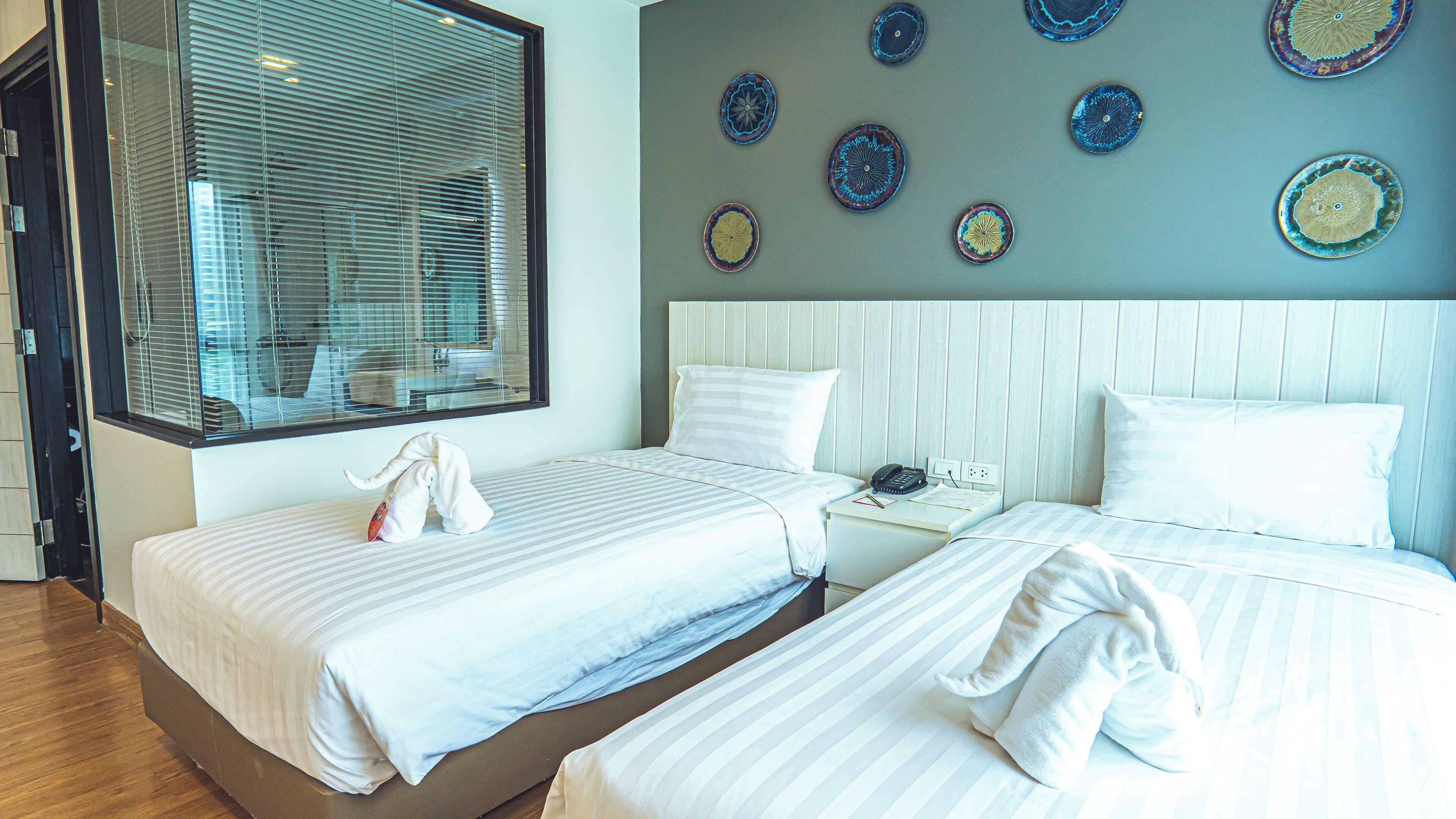 Habitación de hotel con dos camas, un ventanal con persianas, una mesita de noche con teléfono y placas decorativas en la pared.