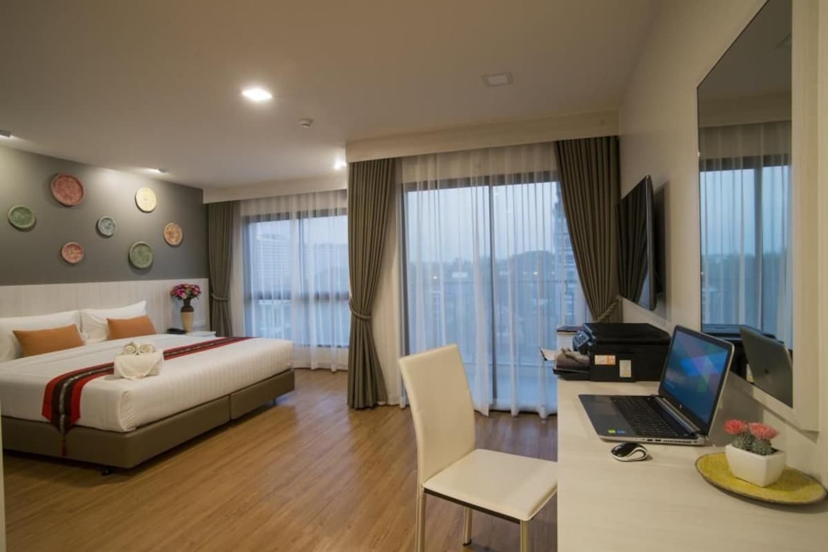 A hotel room with a bed, a desk with a laptop, a chair, and a large window with curtains.