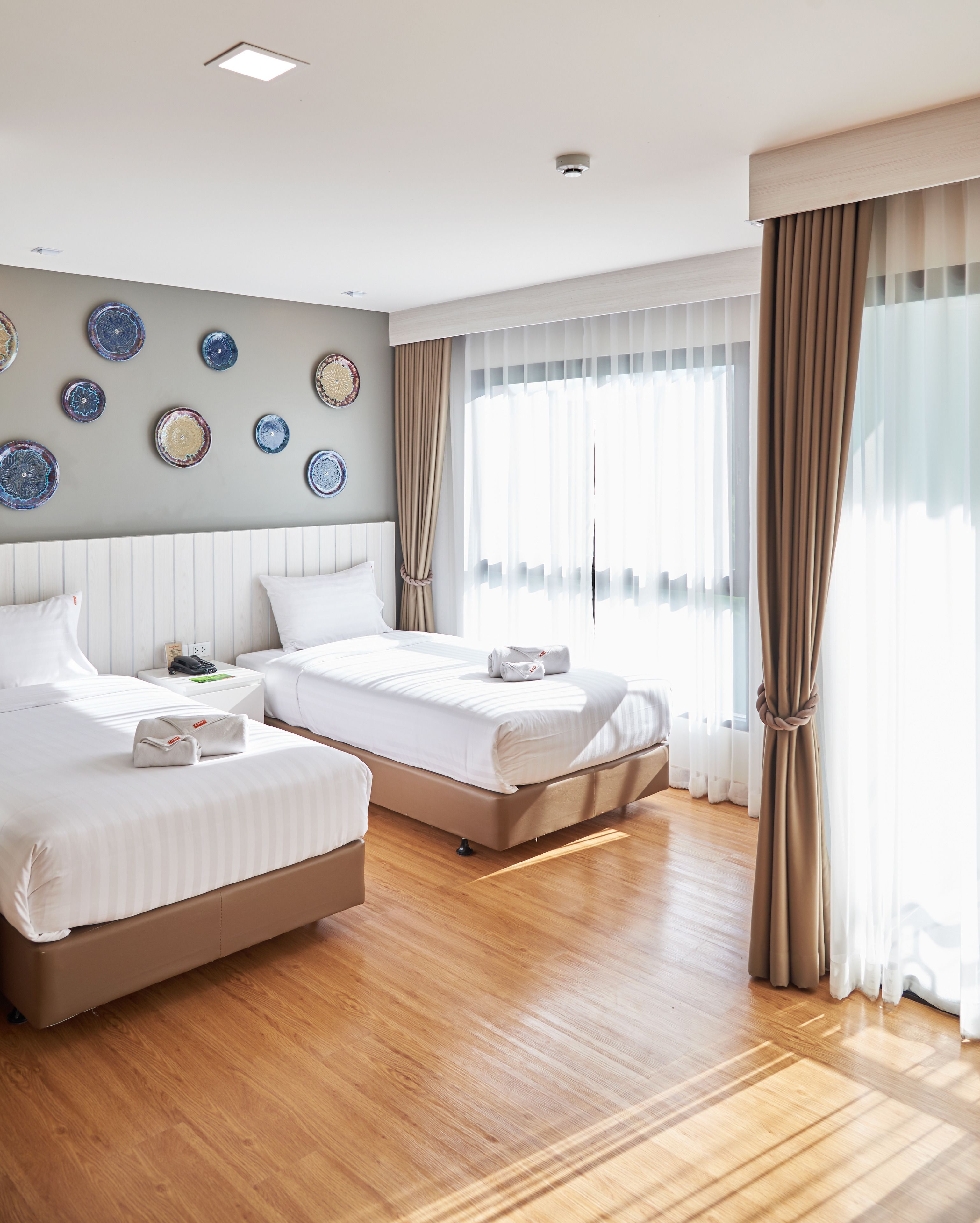 A hotel room with two beds, a wooden floor, a wall with decorative plates, and sheer curtains.