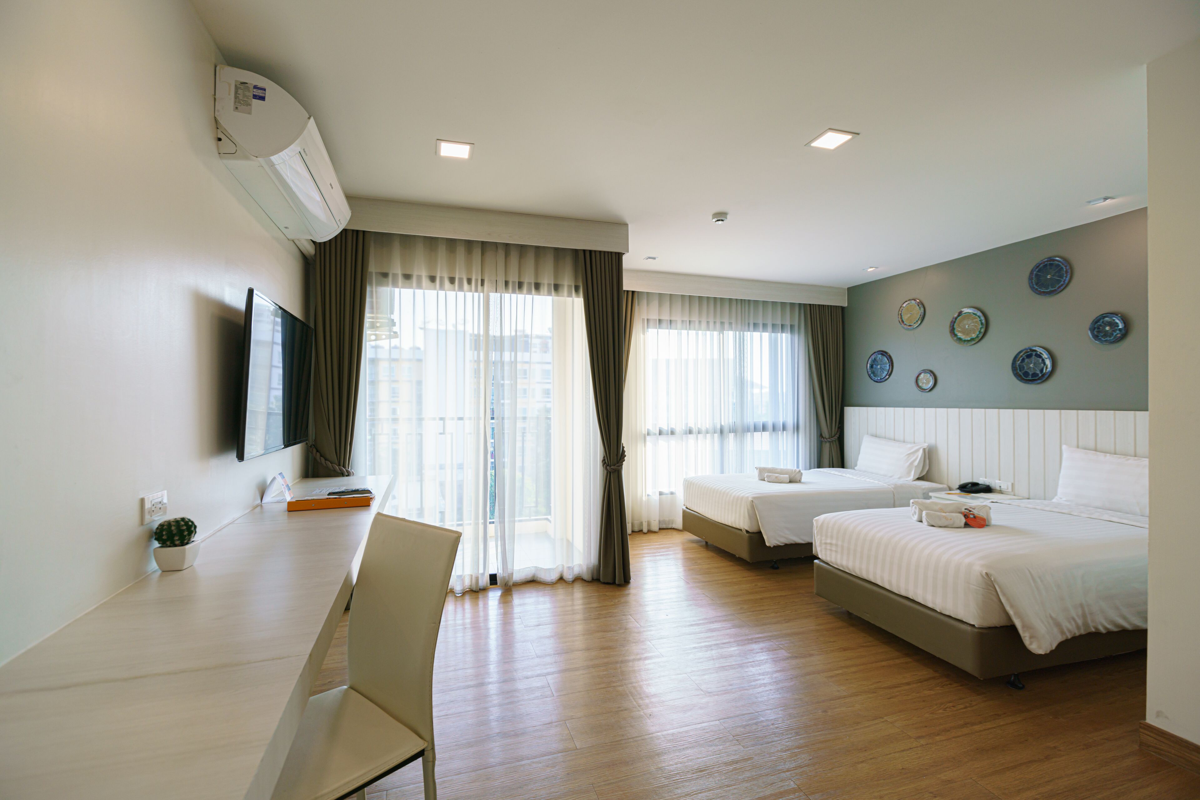 Ein Hotelzimmer mit Bett, Schreibtisch und Stuhl. Ein Fernseher hängt an der Wand, und ein Fenster mit Vorhängen ist vorhanden.