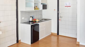 A modern kitchen with white cabinetry, a black dishwasher, and a microwave. There is a sink with a faucet, and a small countertop with various kitchen items. The floor is wooden, and there are two doors visible, one with a sign on it.