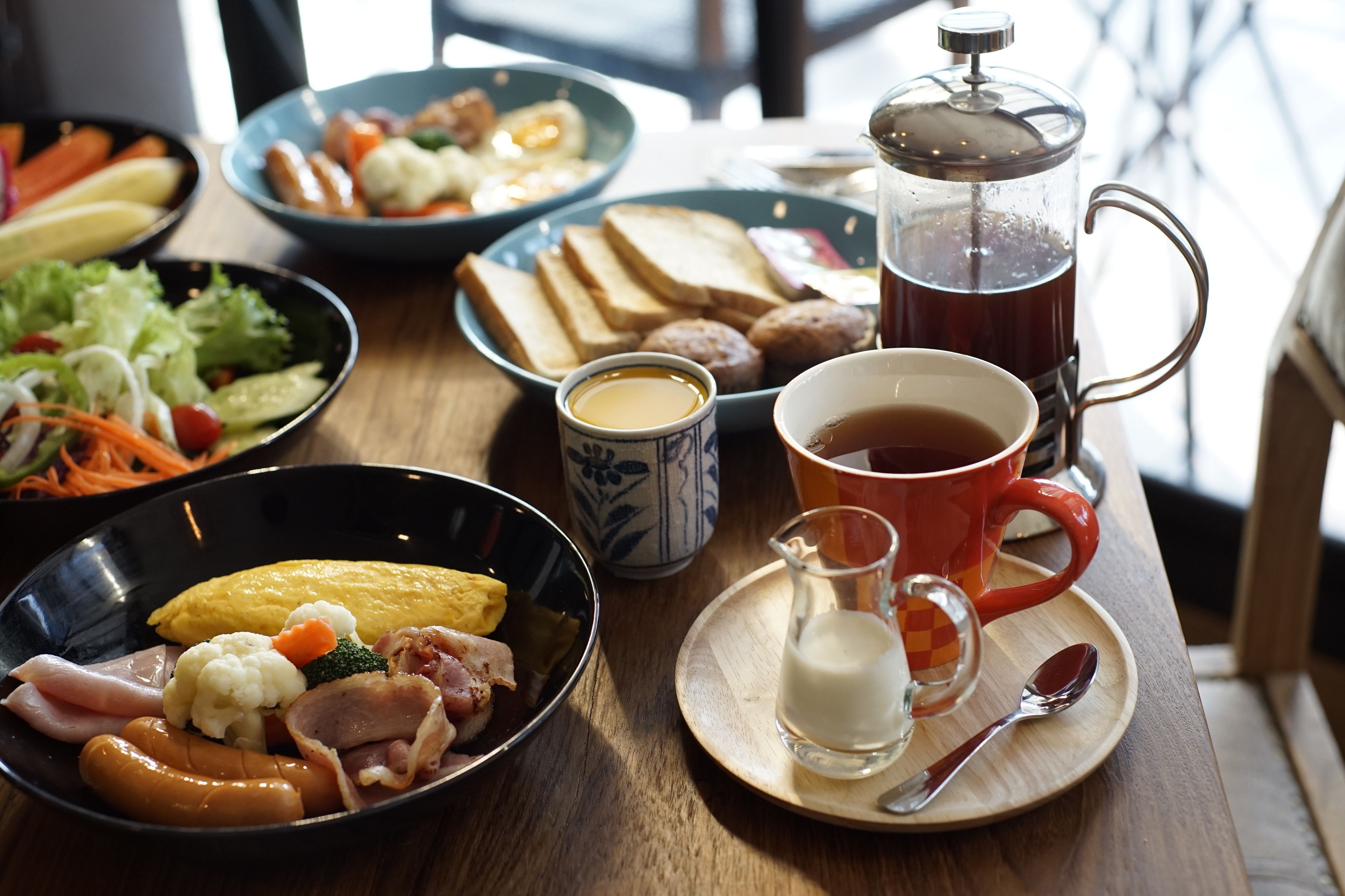 Una mesa de desayuno con diversos platos que incluyen huevos, salchichas y verduras, una cafetera y una taza de café.