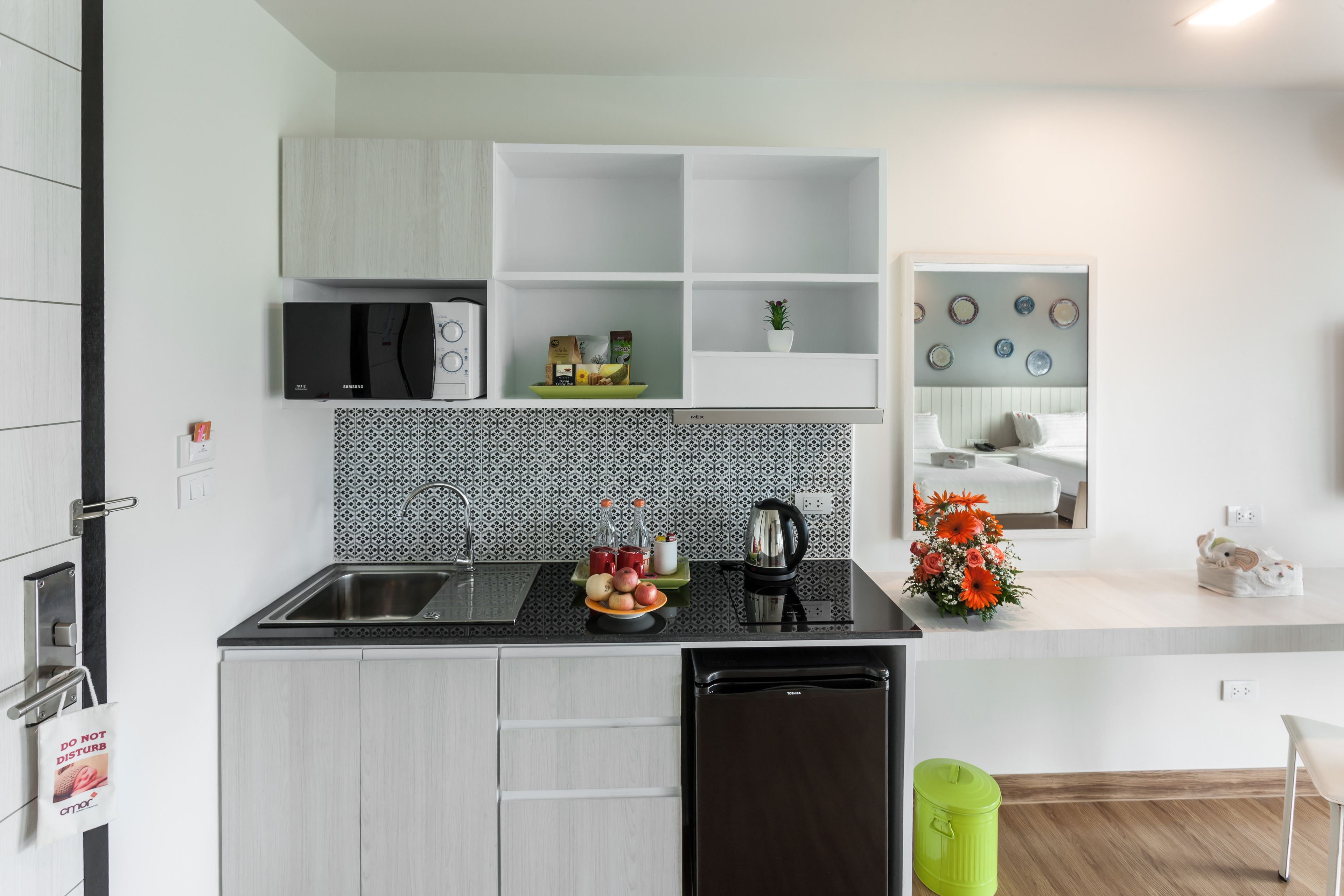 A compact kitchen with a black dishwasher, a sink, and a microwave. There are cabinets above the countertop and a small shelf with decorative items.