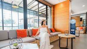 A modern hotel lobby with a large window, a sofa with orange cushions, a wicker table, and a sign indicating cashless payments. - Cmor by Recall Hotels, Chiang Mai (Chiang Mai)