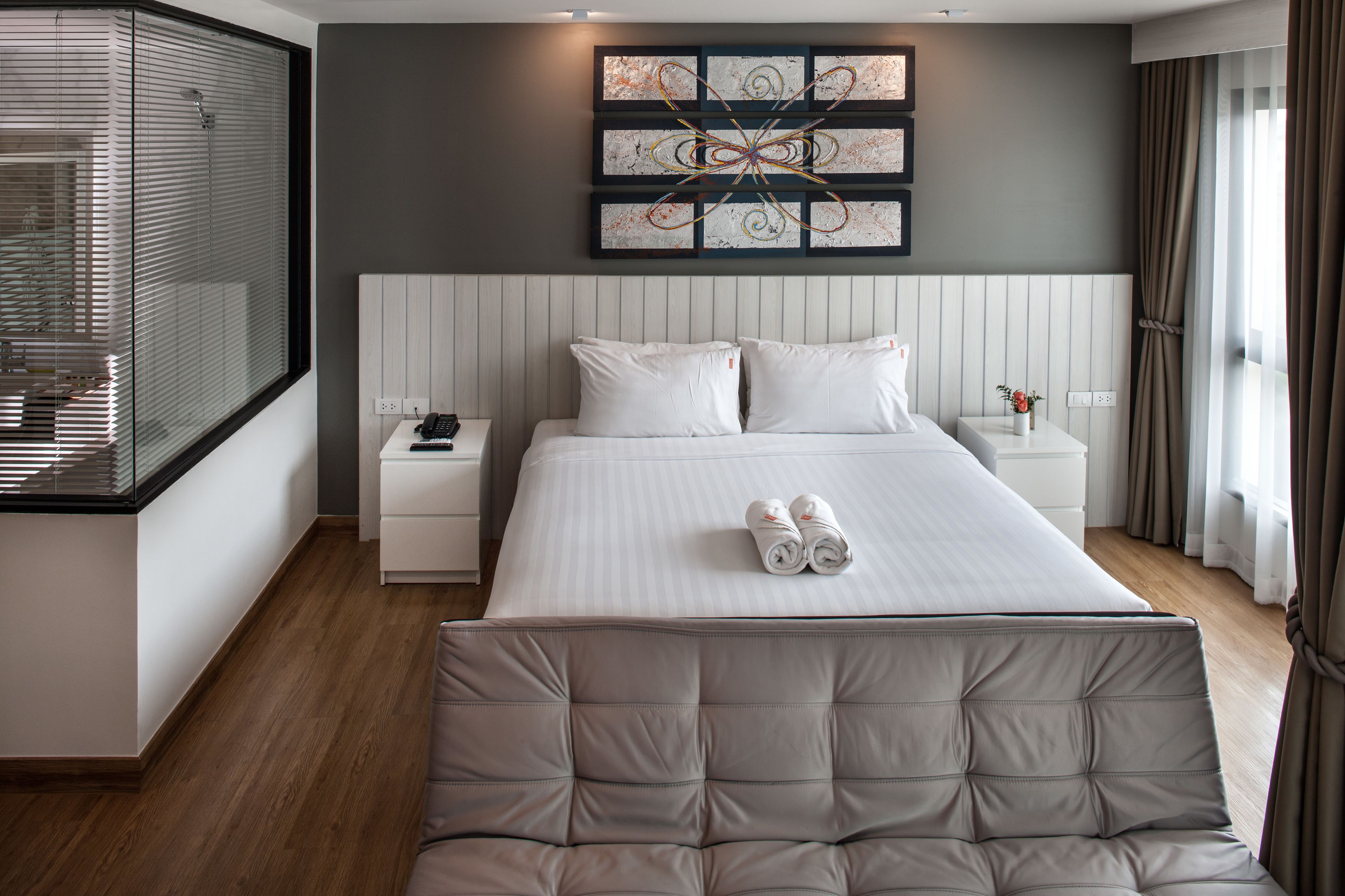 A modern hotel room with a large bed, a bedside table, a nightstand, and a window with blinds.