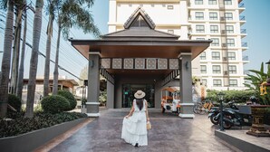 A woman in a white dress walking towards a building with a covered entrance, palm trees, and parked motorcycles. - Cmor by Recall Hotels, Chiang Mai (Chiang Mai)