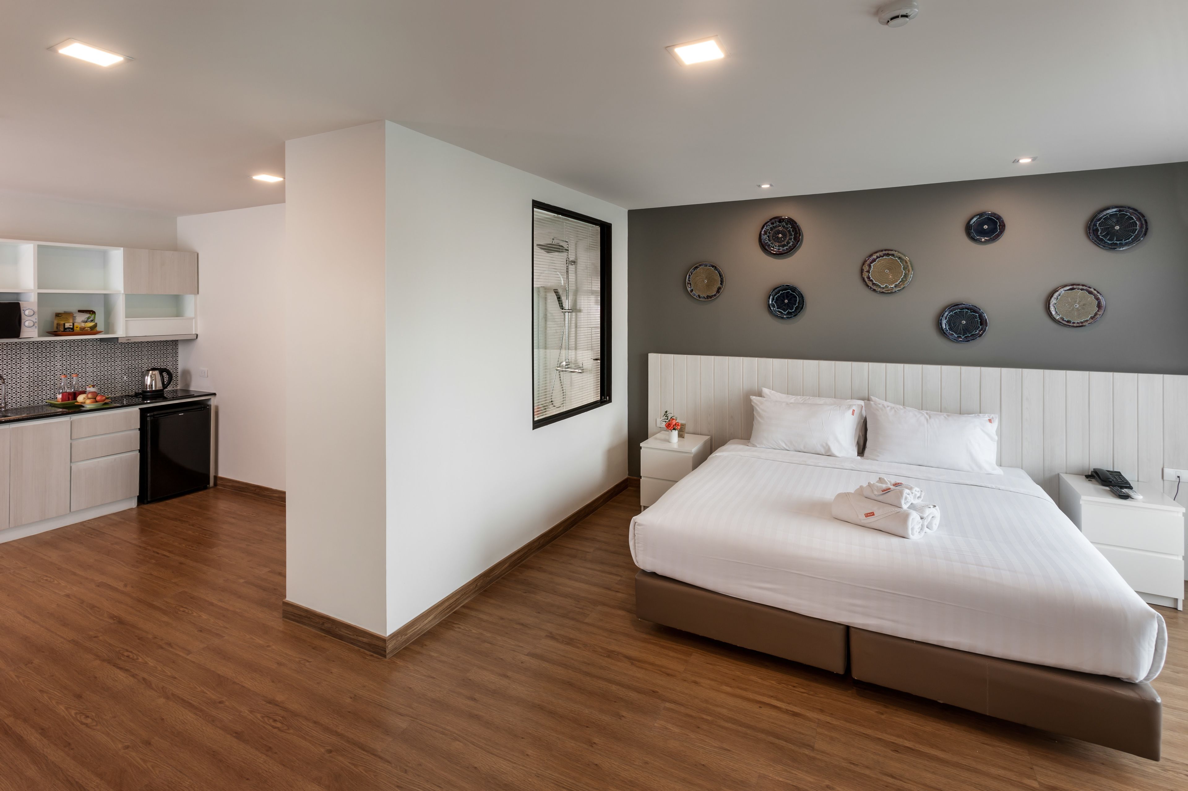 A modern hotel room with a large bed, a small kitchen area, and a bathroom with a glass shower enclosure.
