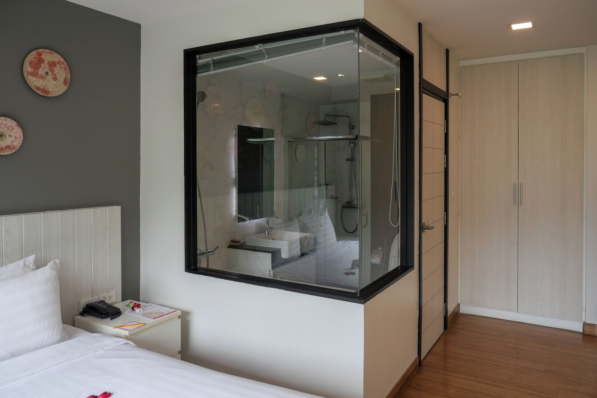 A modern hotel room with a large glass window, a bed, a nightstand, a bathroom with a shower, and a wardrobe.