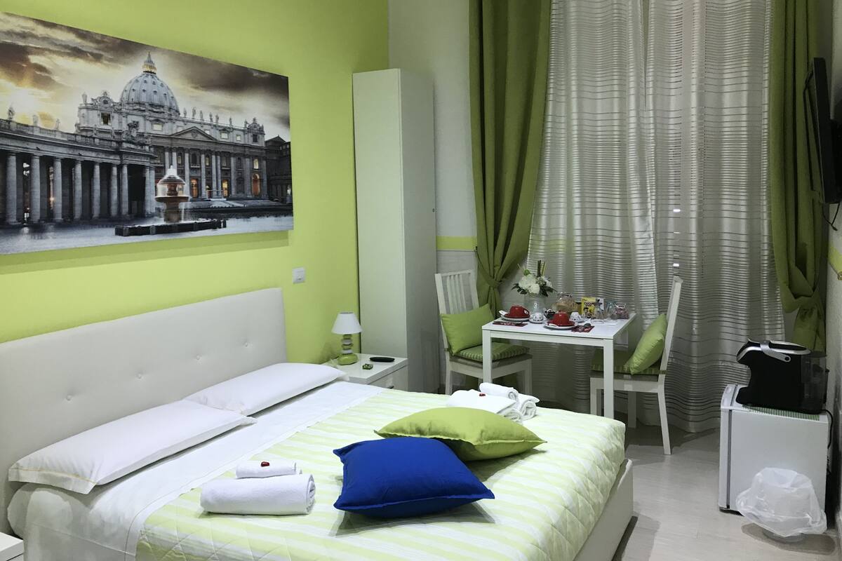 Een hotelkamer met een bed, een tafel met bloemenvaas, een stoel, een bureau met computer, en een schilderij van een historisch gebouw aan de muur.