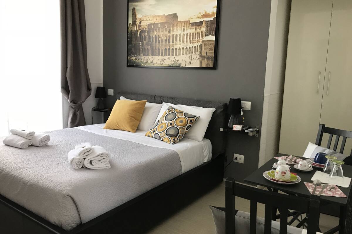 Een netjes opgemaakt bed met handdoeken, een nachtkastje met een lamp, een ingelijste foto aan de muur, en een klein tafeltje met een theepot en kopjes.