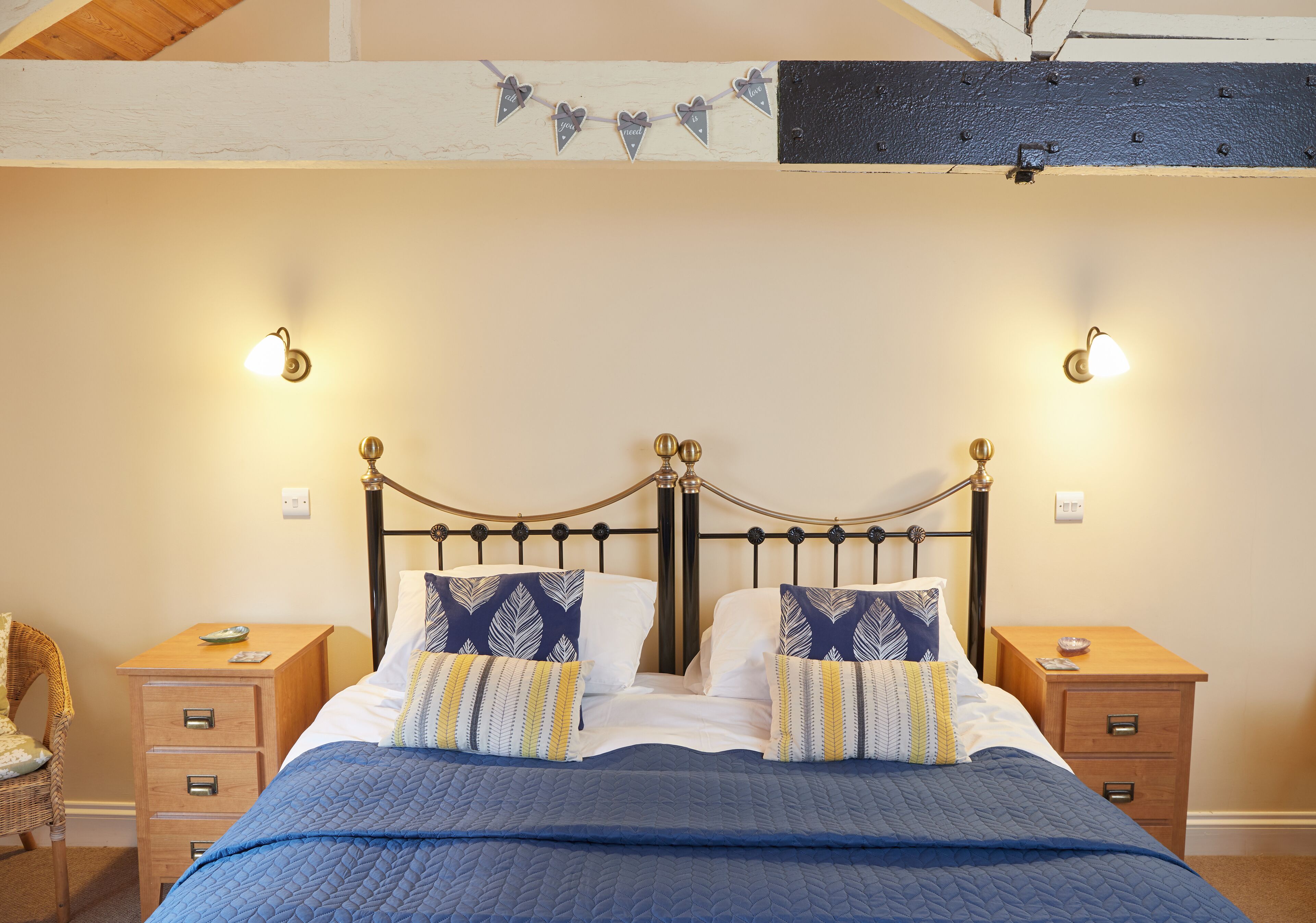 Una cama bien hecha con un edredón azul, dos mesitas de noche con lámparas y un cartel decorativo con forma de corazón en la pared.