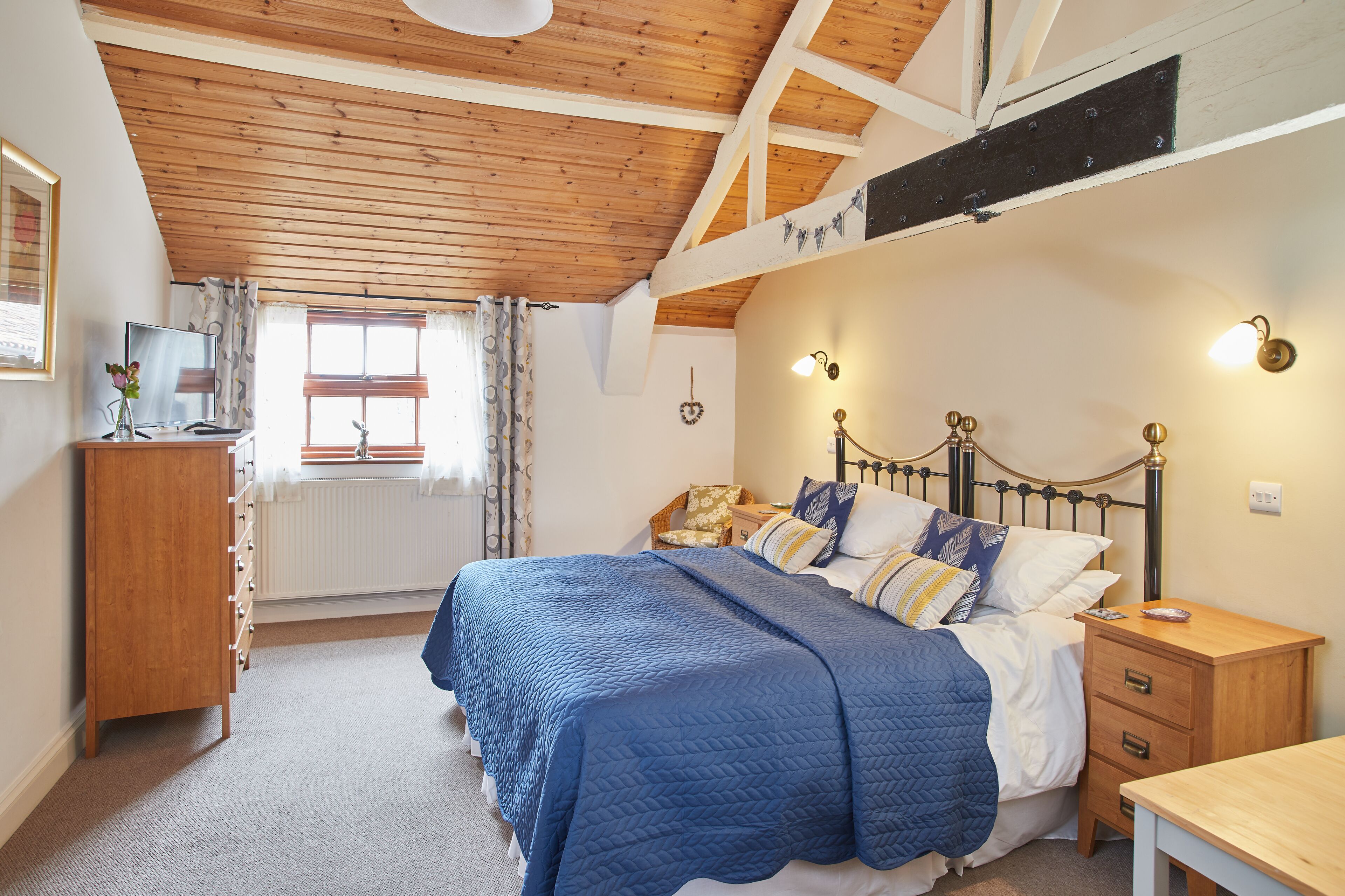 A bedroom with a wooden ceiling, a bed with a blue quilt, a wooden nightstand, and a dresser.