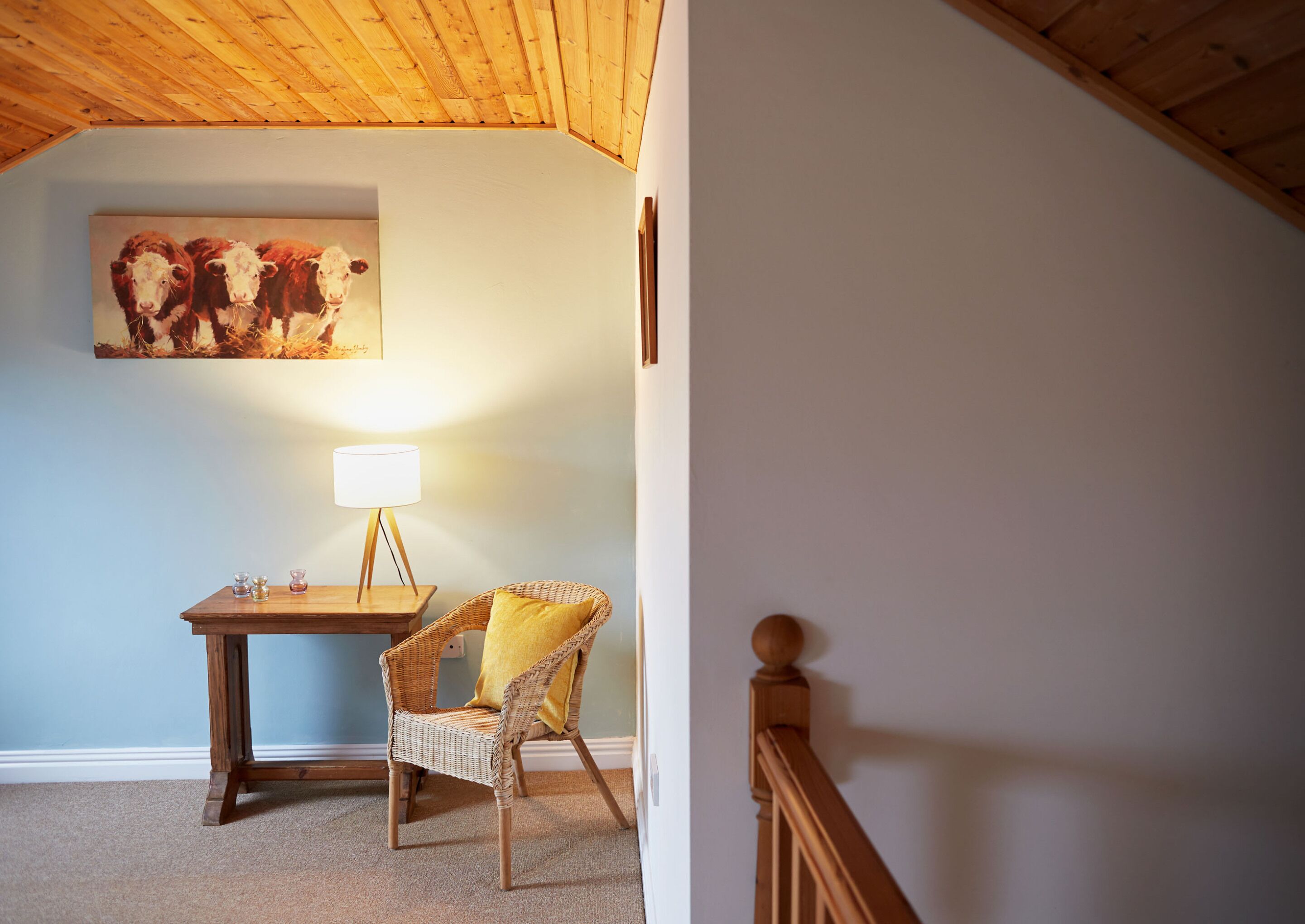 Ein Zimmer mit Holzboden, einem Kuhbild, einem Tisch mit Lampe, einem Korbstuhl und einer Treppe.