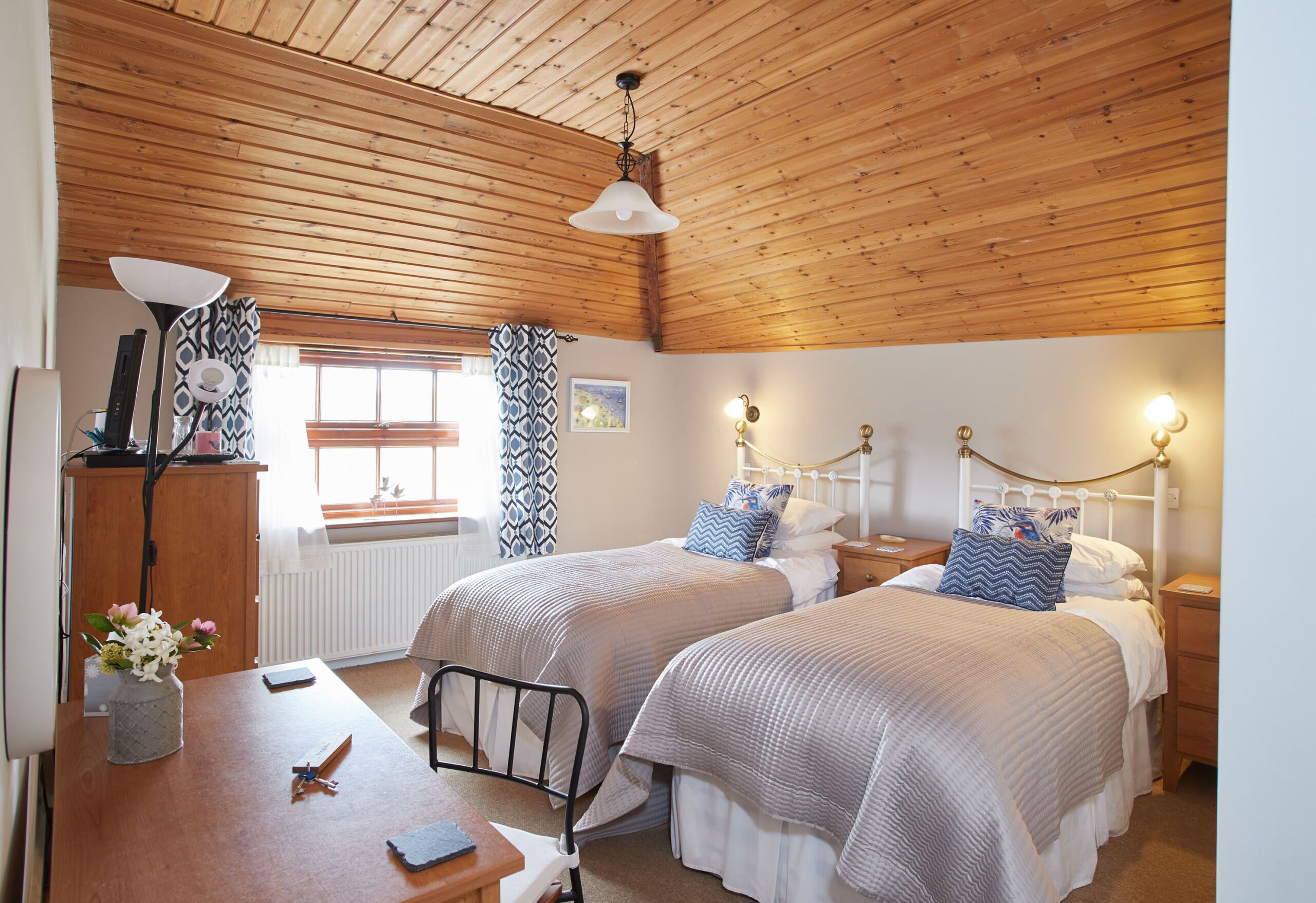 Une chambre avec deux lits, un plafond en bois, une table ornée d’un vase de fleurs et une fenêtre avec des rideaux à motifs.