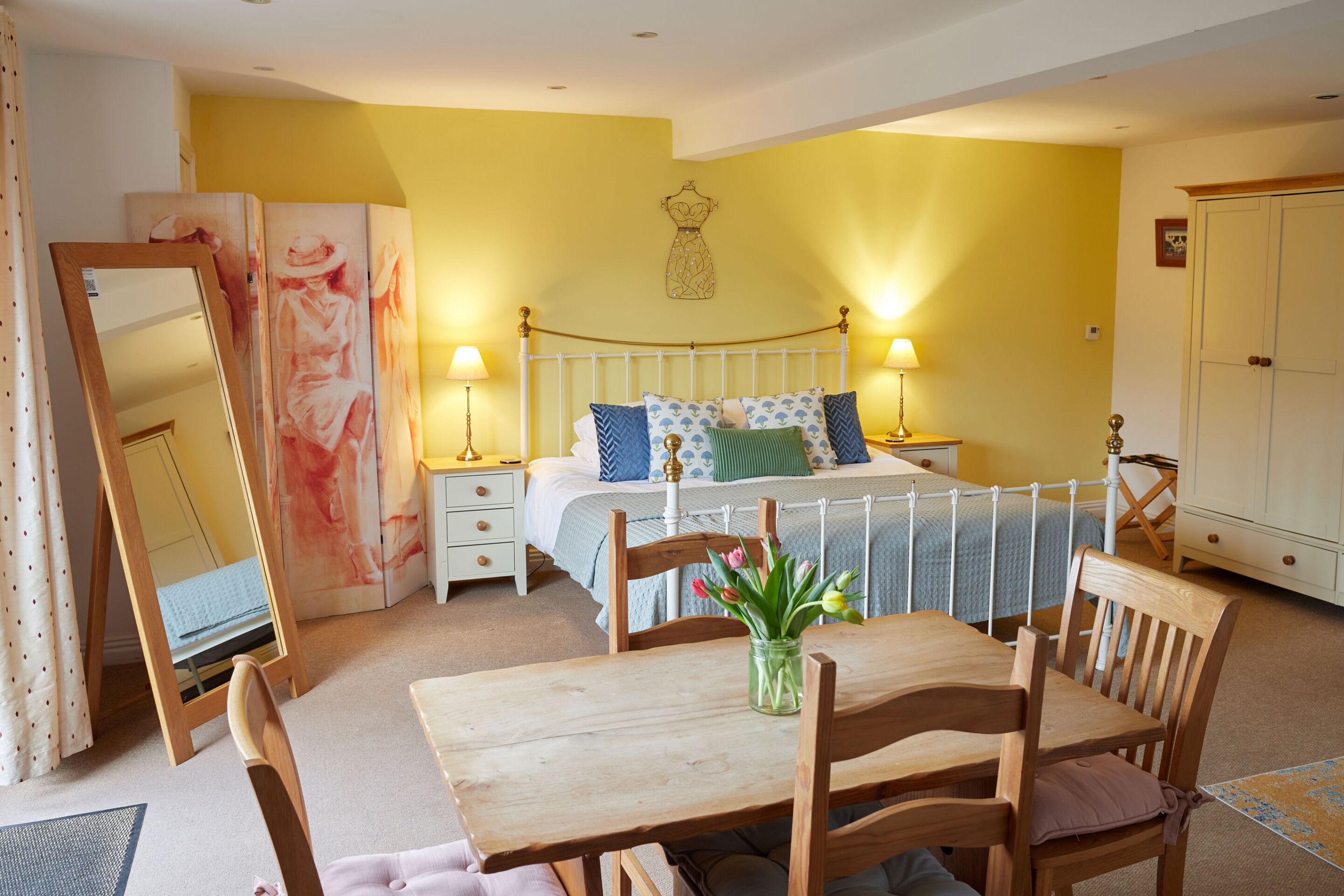 A bedroom with a bed, bedside tables, a mirror, a wooden table, chairs, and a vase with flowers.