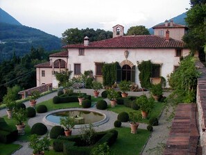 Una villa con jardín que presenta un estanque circular, setos perfectamente podados y plantas en macetas.