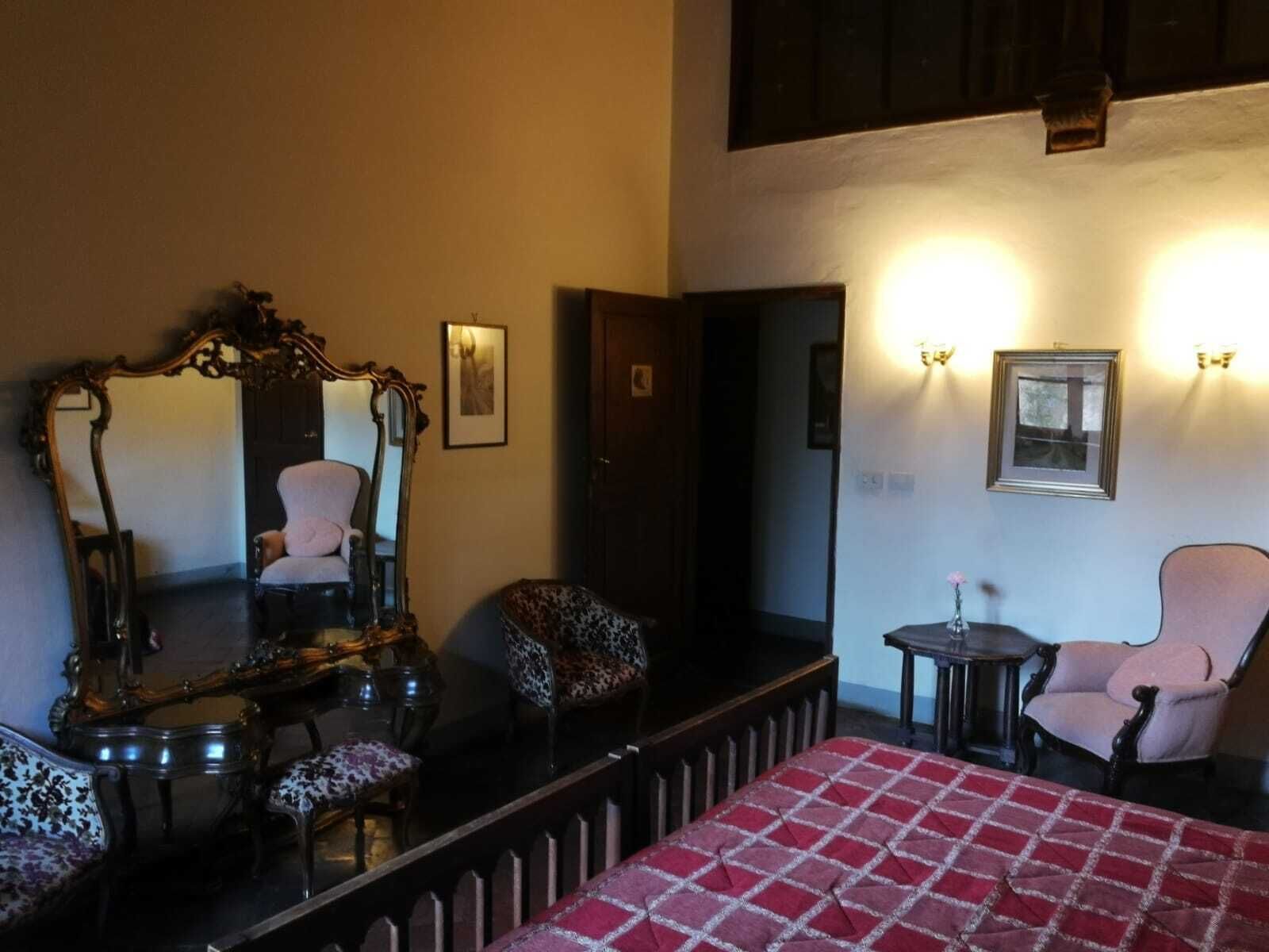 Habitación de hotel con un espejo grande, una cama con colcha a cuadros blancos y rojos, dos sillones, una mesita con un jarrón y luces empotradas en la pared.