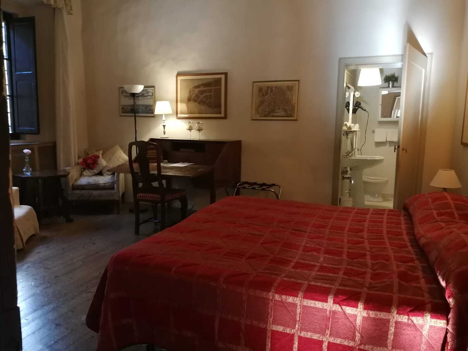 A hotel room with a large bed, a desk with a chair, a small table with a vase, and a door leading to a bathroom.
