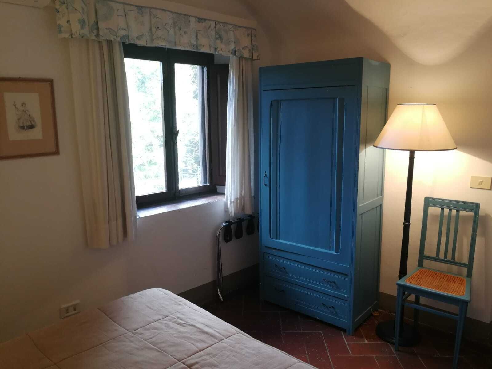 A bedroom with a bed, a window with curtains, a blue wardrobe, a chair, and a floor lamp.