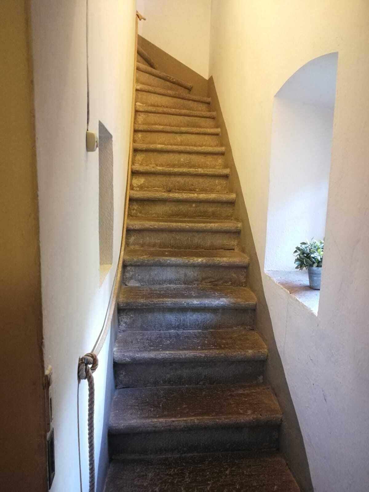 Un escalier étroit avec des marches en pierre menant vers le haut, bordé de murs sobres et surmonté d’une petite fenêtre.