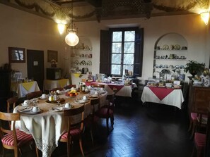 A dining area with multiple tables set for meals, surrounded by chairs. There are hanging light fixtures, a window with shutters, and shelves with decorative items.