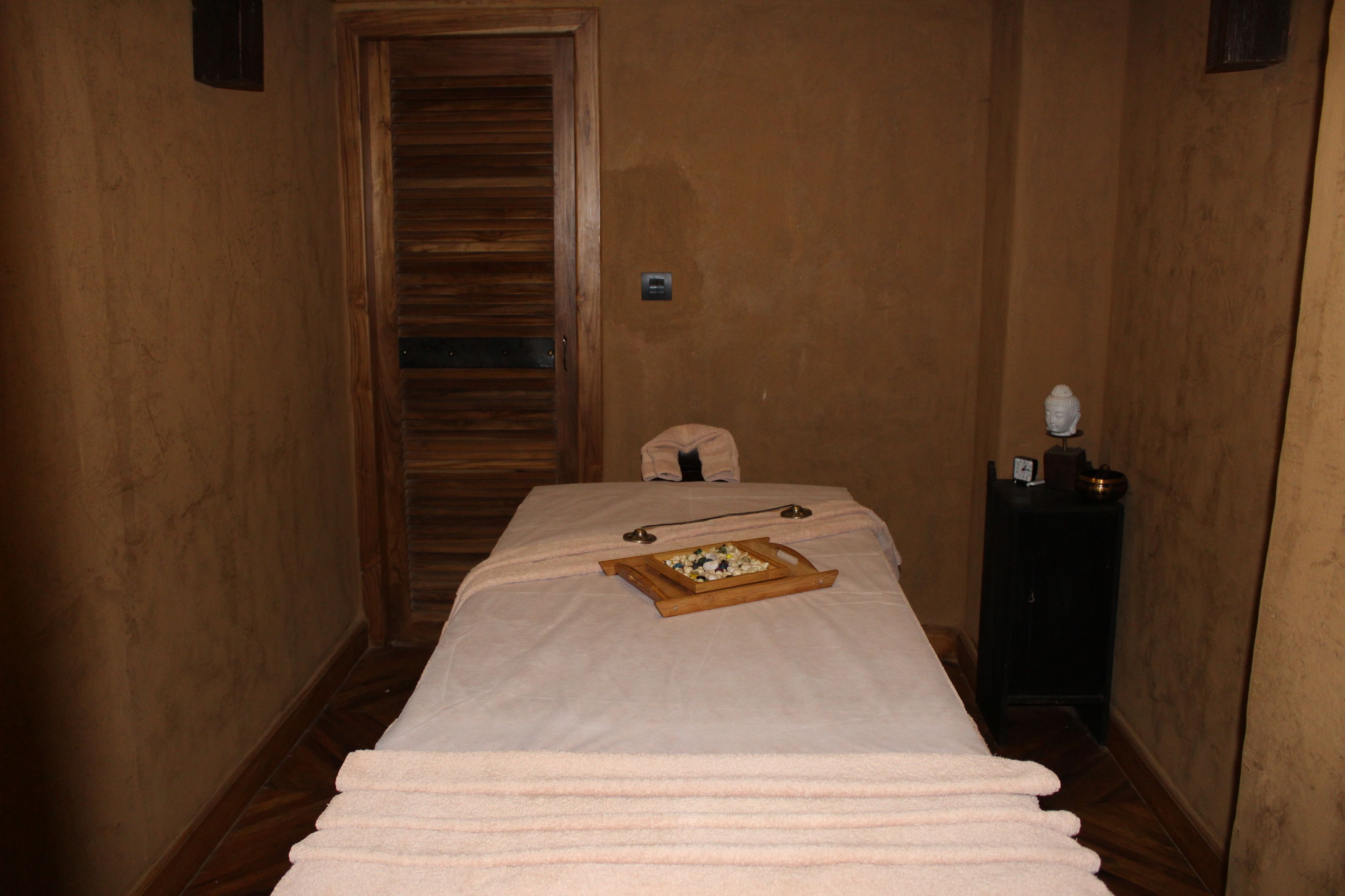 Una habitación con puerta de madera, cama con manta blanca, mesita de noche con lámpara y bandeja con comida.