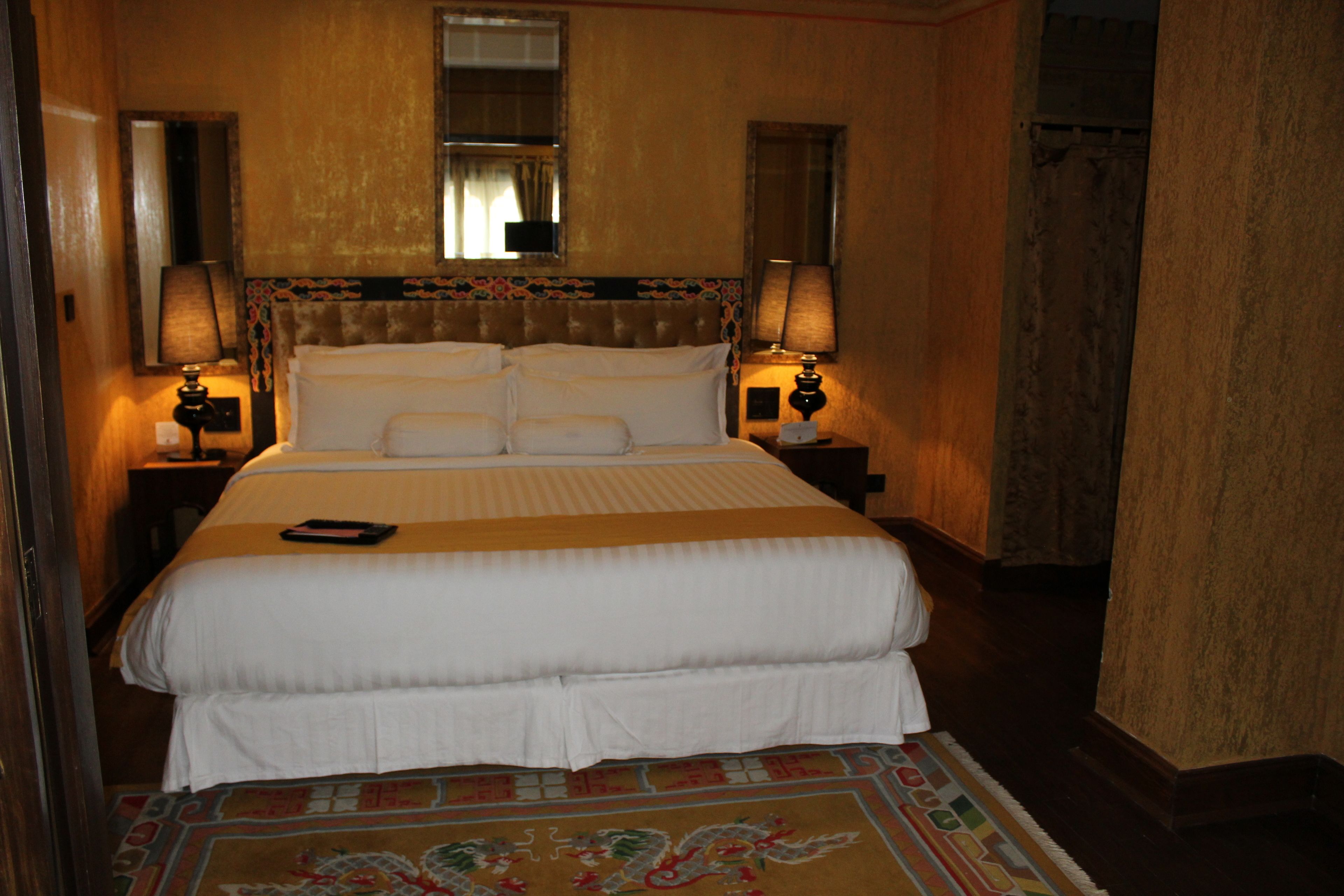 A hotel room with a large bed, two bedside tables with lamps, a window with curtains, and a patterned carpet.