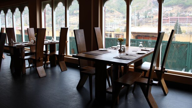 A dining area with wooden tables and chairs, large arched windows, and a view of a cityscape. - Druk Hotel (Thimphu)
