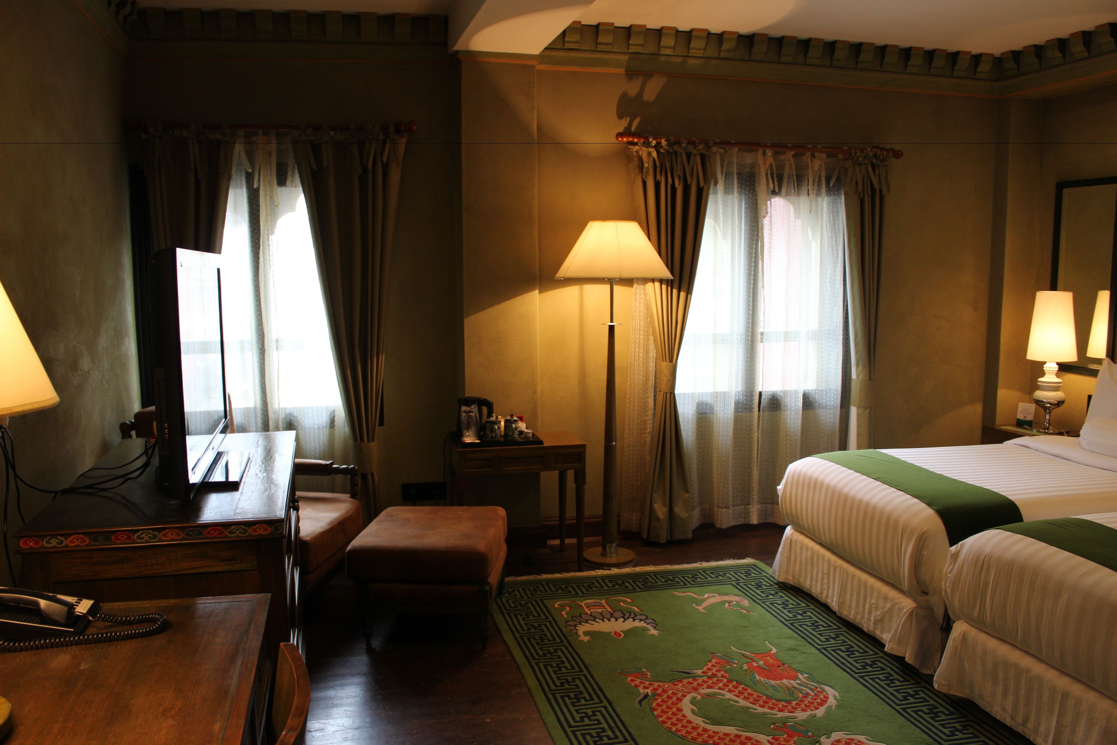 A hotel room with a bed, two bedside tables, a desk with a telephone, a chair, a lamp, and a decorative rug.