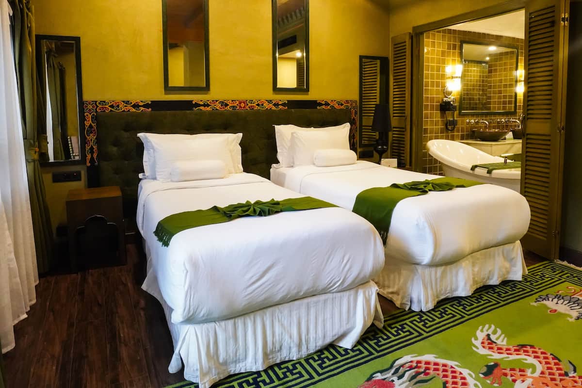 A hotel room with two beds, a large mirror, a bathroom with a bathtub, and a green patterned rug.