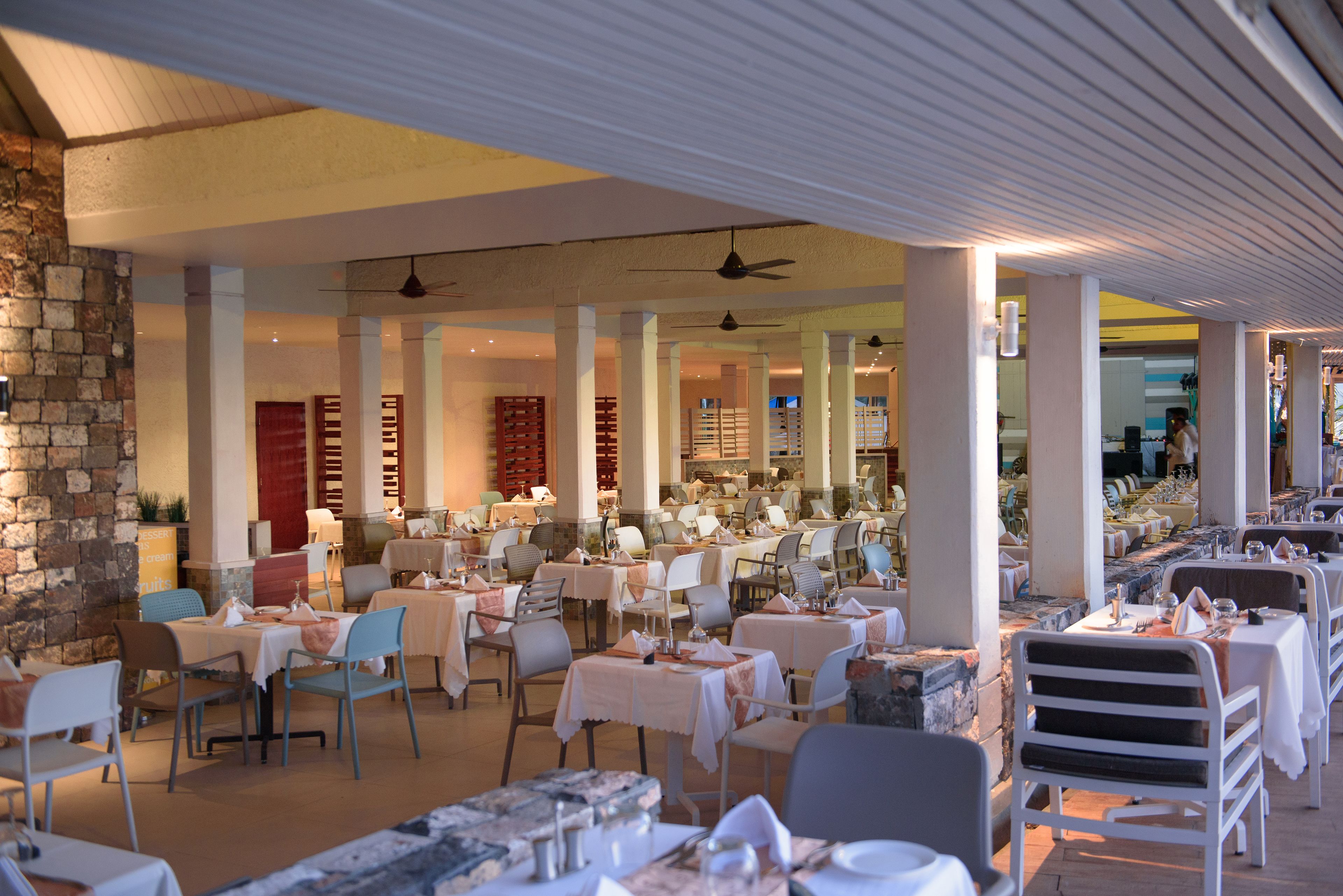 Un restaurante con varias mesas y sillas, una pared de piedra en un lateral y ventiladores de techo.