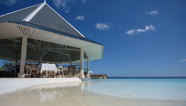 Un pavillon situé au bord de la plage, offrant une vue dégagée sur l’océan, avec un mur en pierre et une table blanche entourée de chaises.