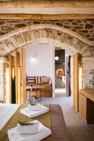Una habitación acogedora con un arco de piedra, una cama con cabecera verde, un banco de madera y una mesita con un jarrón de flores.