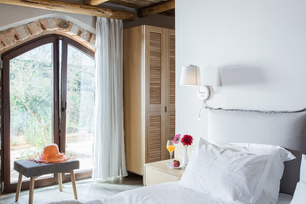 Une chambre d’hôtel avec un plafond en bois, un lit recouvert de draps blancs, une petite table avec une serviette, une table de chevet avec une lampe, et une vue sur la verdure par la porte.