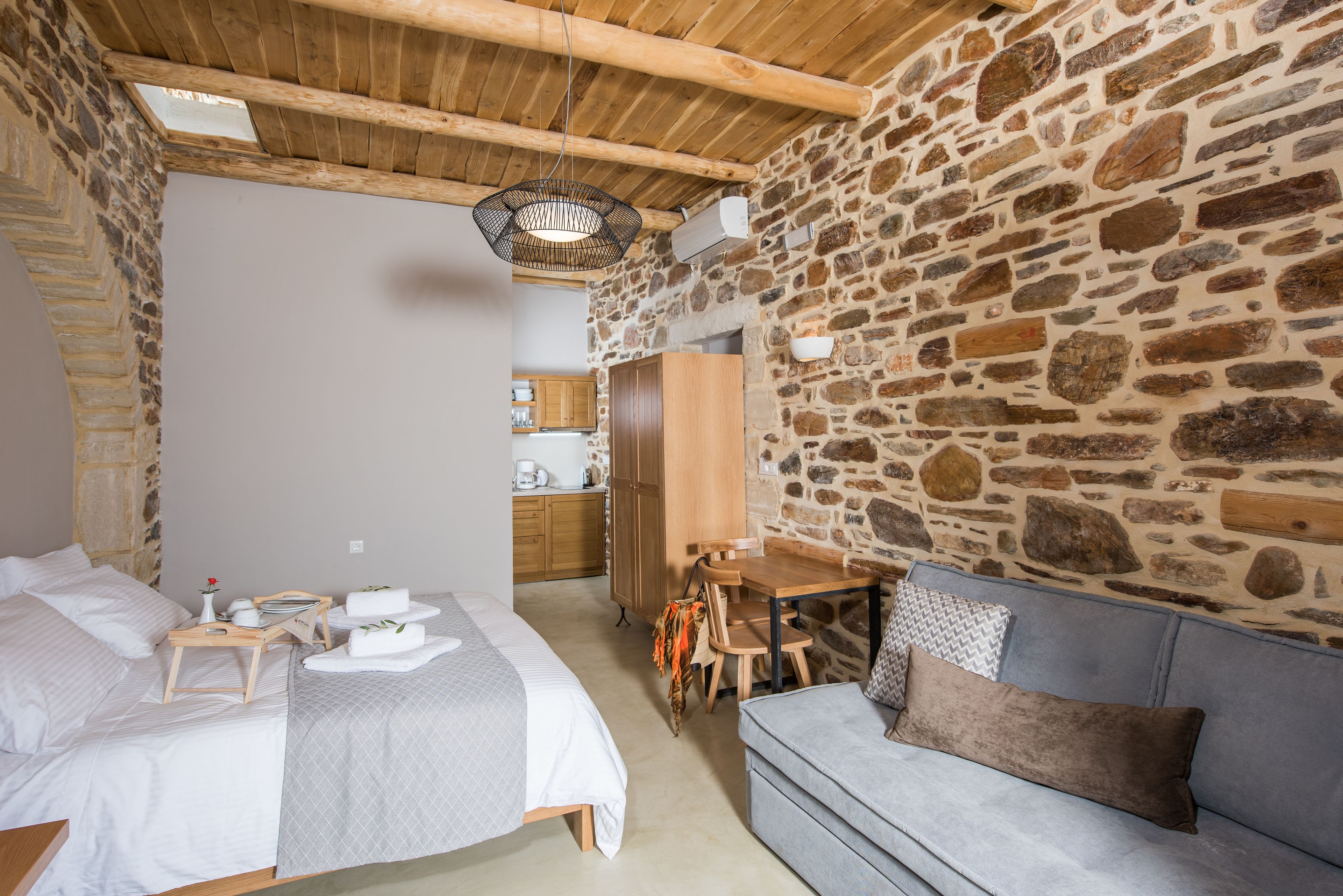 A cozy room with a stone wall, a wooden ceiling, a bed with a wooden nightstand, a gray sofa, and a small wooden table with chairs.