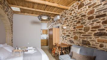 A cozy room with a stone wall, a wooden ceiling, a bed with a wooden nightstand, a gray sofa, and a small wooden table with chairs.