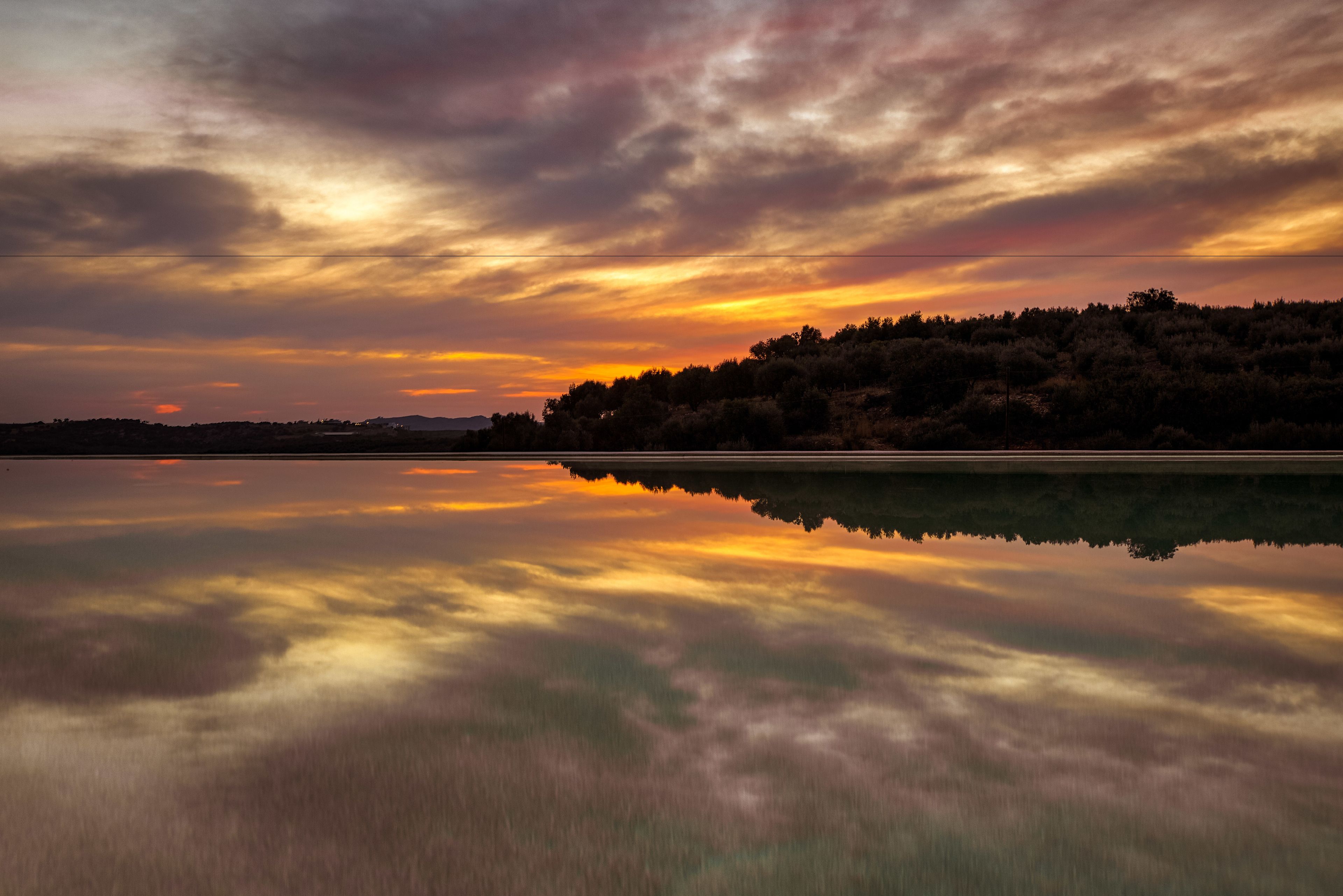 A calm lake reflecting a sunset with a forested hill in the background.